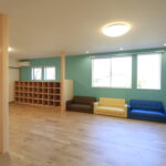 木の温もりあふれる新築学童施設の内観。アクセントカラーのティールブルーの壁、造り付けの木製ロッカー、カラフルなミニソファが並ぶプレイルーム。